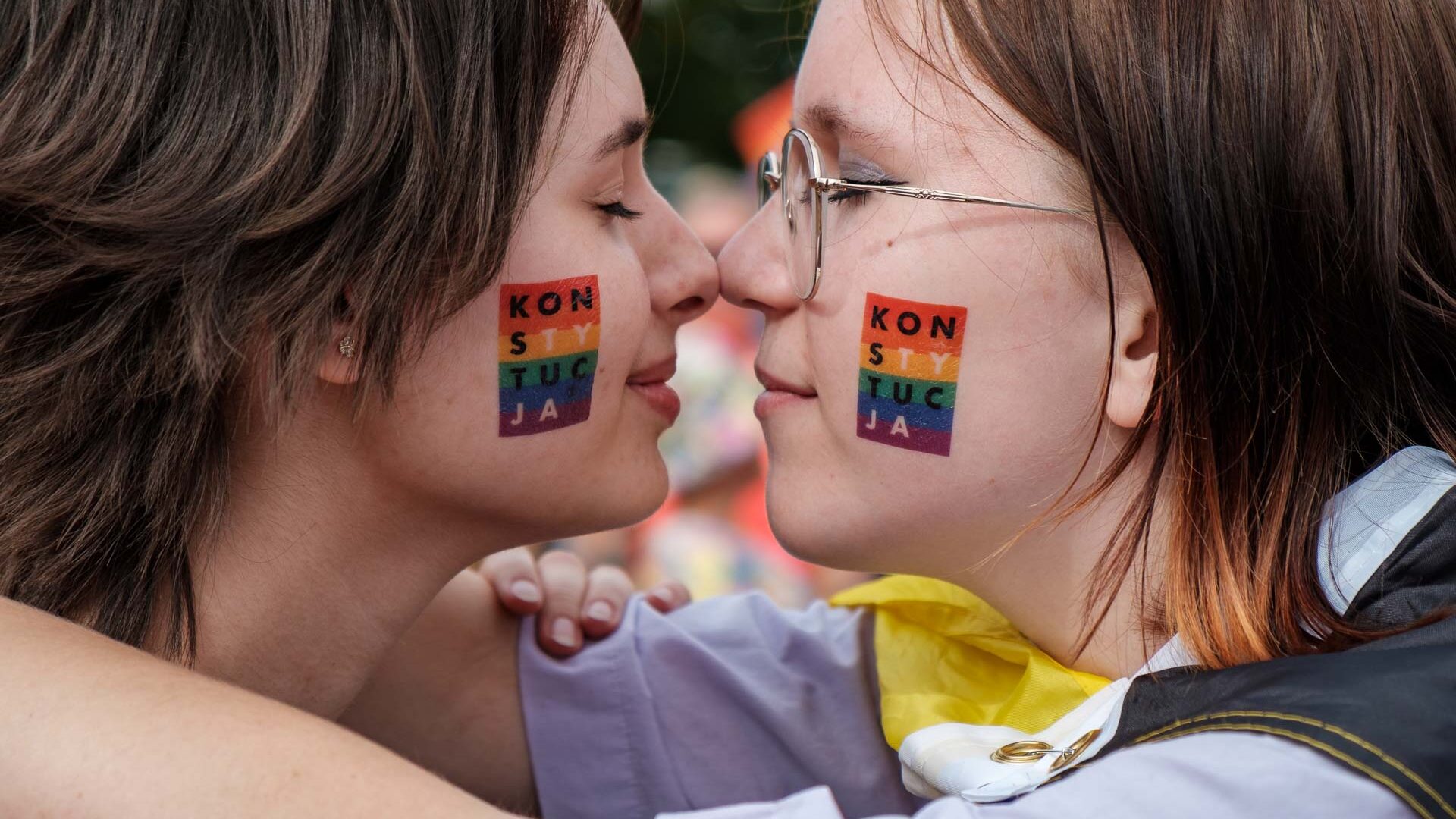 Dwie dziewczyny stykające się nosami z wymalowanymi flagami LGBT na policzkach i wpisanym w nie słowem KONSTYTUCJA